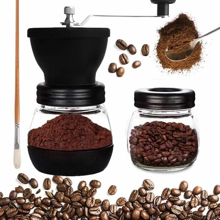 Manual coffee grinder with a clear glass jar and black top, shown alongside a small glass bean jar, loose coffee beans, ground coffee, a spoon scoop, and a wooden-handled brush on a white background.