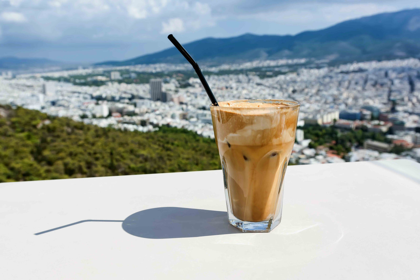 Iced Greek Nescafé frappé served in a tall glass with a black straw, set against a scenic backdrop of a sunny cityscape and mountains.