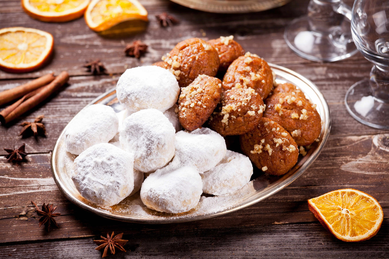 A plate of traditional Greek cookies featuring powdered sugar–coated kourabiedes and honey-drenched melomakarona topped with crushed walnuts, displayed on a rustic wooden table with cinnamon sticks, star anise, and dried orange slices.