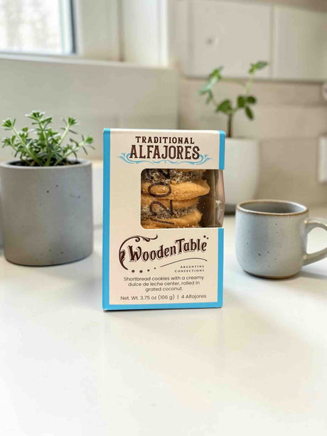 A box of WoodenTable Traditional Alfajores cookies sits on a white countertop in a bright kitchen setting, flanked by a gray ceramic mug and potted green plants.