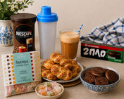 Mediterranean-inspired Greek frappé bundle featuring NESCAFÉ Classic instant coffee, a blue frappé shaker bottle, assorted loukoumi delights, traditional Greek koulourakia cookies, chocolate sandwich cookies in a decorative bowl, and a freshly prepared iced frappé drink styled on a warm kitchen countertop with blue-and-white accents.