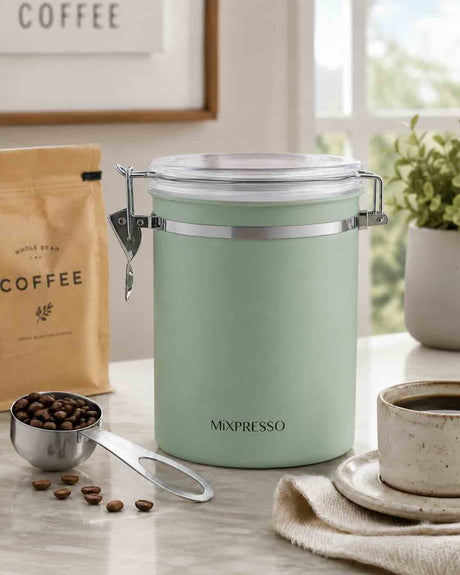A mint green metal coffee storage jar with a clear clamp-lid container, shown on a marble countertop with a coffee bean bag, measuring cup, coffee beans, and a cup of coffee in a bright kitchen setting.
