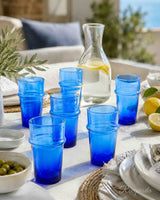 A set of cobalt blue stacked drinking glasses arranged on a styled outdoor dining table, alongside a glass carafe, lemons, olives, plates, and a blurred patio background.