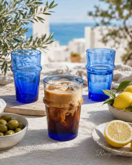 A set of cobalt blue stacked drinking glasses arranged on a light stone table outdoors, with an iced coffee in the center glass, lemons, olives, and a blurred coastal background.