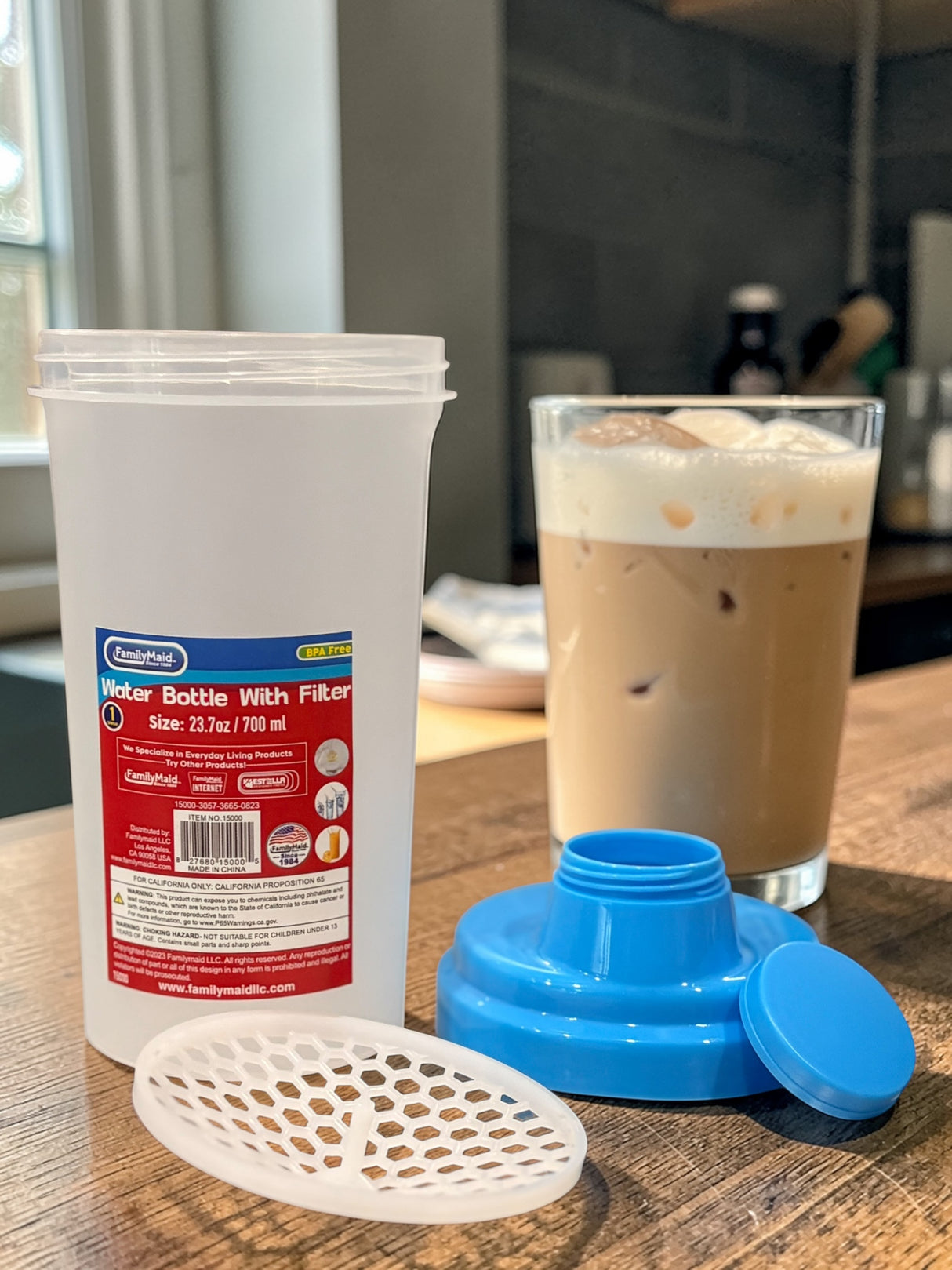 White container with red label, blue lid, and white filter on a wooden surface with a glass of iced coffee.