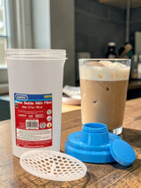 White container with red label, blue lid, and white filter on a wooden surface with a glass of iced coffee.