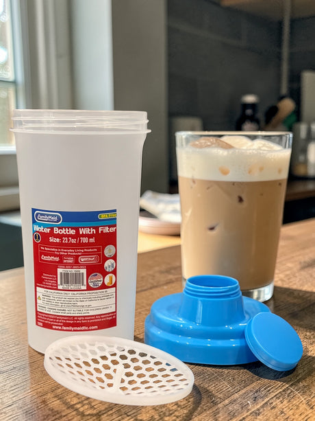 White container with red label, blue lid, and white filter on a wooden surface with a glass of iced coffee.