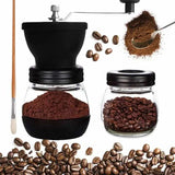 Manual coffee grinder with a clear glass jar and black top, shown alongside a small glass bean jar, loose coffee beans, ground coffee, a spoon scoop, and a wooden-handled brush on a white background.