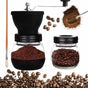 Manual coffee grinder with a clear glass jar and black top, shown alongside a small glass bean jar, loose coffee beans, ground coffee, a spoon scoop, and a wooden-handled brush on a white background.