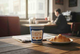 A blue and white paper coffee cup with the text 'WE ARE HAPPY TO SERVE YOU' sits on a wooden table in a cafe setting, next to a croissant on a plate and a newspaper. A blurred person is seated in the background near a window.